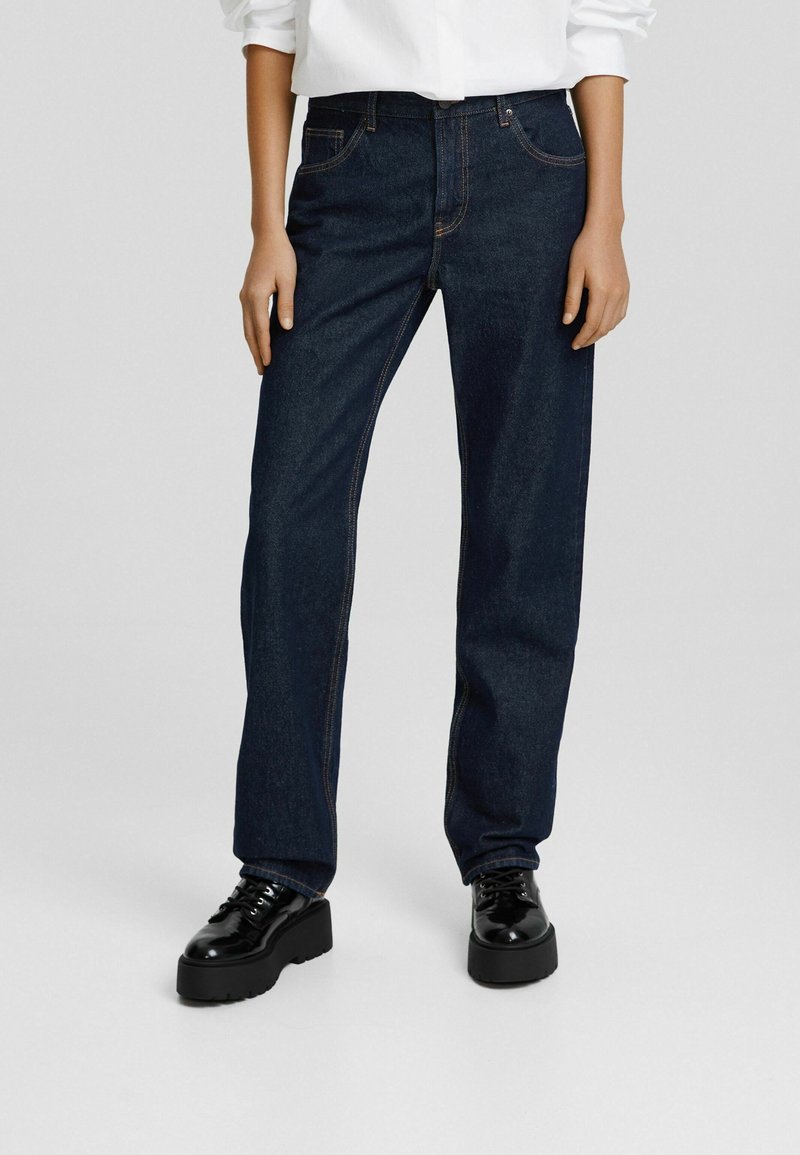 Jeans de denim azul oscuro con un corte recto, con costuras naranjas, cinco bolsillos y una cintura estándar. Llevados con zapatos de plataforma negros.