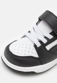 Baskets noir et blanc avec un dessus en cuir, matériaux texturés, perforations au niveau des orteils et une bande Velcro sur les lacets.
