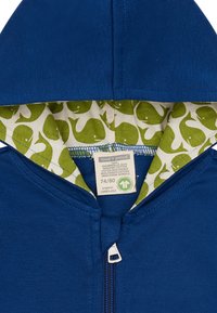 Felpa blu con zip realizzata in cotone biologico, con fodera interna a motivo balena verde e un'etichetta che mostra la taglia e le istruzioni per la cura.
