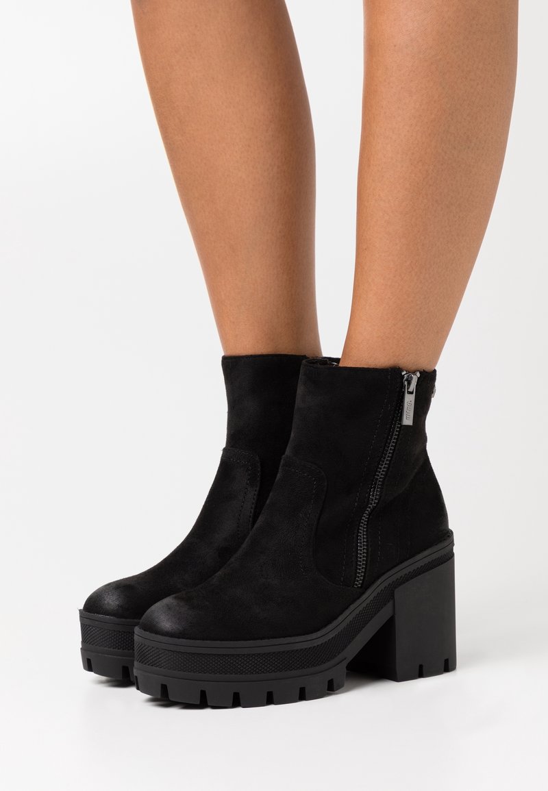 Bottines en daim noir avec talons carrés épais et fermetures éclair latérales portées sur des jambes nues sur fond blanc.