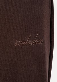 Σκούρο καφέ ριγωτό ύφασμα με το λογότυπο «smilodox» κεντημένο με κλωστή στο ίδιο καφέ χρώμα σε γραφικό στυλ καλλιγραφίας.