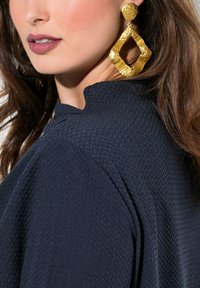 Top navy texturizzato con colletto alto; grandi orecchini dorati con forma geometrica e dettagli intricati completano l'outfit.
