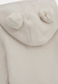Haut à capuche en tricot crème avec une surface texturée, présentant deux accents en pompons en peluche sur la capuche et un design simple et décontracté.