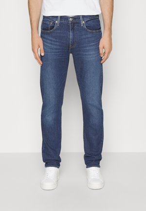 Mann trägt dunkelblaue Slim-Fit-Jeans und weiße Turnschuhe und steht vor einem schlichten hellen Hintergrund.