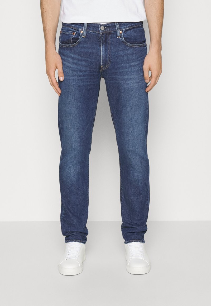 Homme portant un jean slim bleu foncé et des baskets blanches debout devant un fond clair uni.