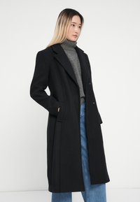 Cappotto lungo nero con design su misura, caratterizzato da un ampio bavero, due bottoni e tasche laterali, indossato sopra un dolcevita grigio e jeans blu.