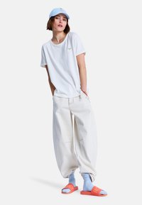 Witte katoenen T-shirt, losse crèmekleurige broek, blauwe sokken en oranje slip-ons met logo. Blauwe pet. Ontspannen pasvorm, casual stijl.