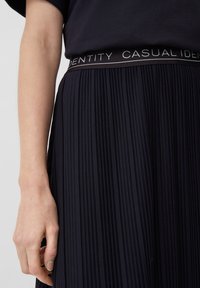 Donna che indossa una gonna pieghettata nera con una fascia etichettata "CASUAL IDENTITY" e una maglietta nera, con la mano appoggiata di lato.
