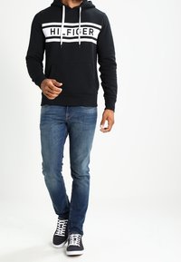 Felpa nera con testo bianco "HILFIGER", tasca a marsupio, cordino regolabile. Indossata con jeans blu e sneakers nere con lacci bianchi.