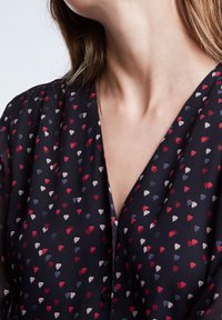 Zwarte blouse met een V-halsontwerp, versierd met kleine rode, blauwe en crème hartpatronen, gemaakt van een lichtgewicht stof.