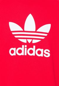 Sudadera roja con un gran logo blanco de Adidas. El logo incluye tres rayas y un diseño de trébol. Tela suave y texturizada.