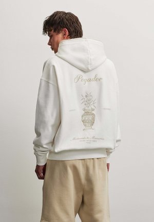 Ung mand iført hvid hættetrøje med blomster-vasedesign og teksten "Pegador" samt beige bukser vender ryggen til mod en ensfarvet baggrund.