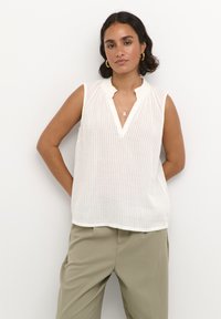 Haut sans manches blanc avec un motif rayé texturé, un col volanté et un décolleté en V. Associé à un pantalon vert olive.