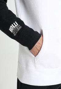 Svartvit hoodie med känguruficka, ribbad mudd och "HOLLISTER"-text på den svarta ärmen. Blandning av bomullsmaterial.