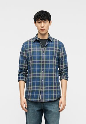 Fiatal férfi rövid fekete hajjal, kék-zöld kockás ingben, fekete póló és kék farmer nadrág fölött, szemben állva.