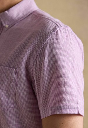 Krótka koszula w kolorze lawendowym w kratę z kieszonką z przodu, noszona przez osobę na jednolitym tle.