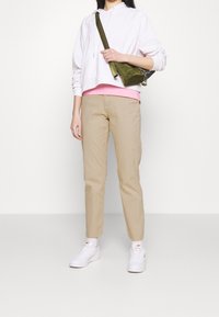 Vit croppad hoodie, rosa lager under, beige taperade byxor, vita sneakers och en olivgrön crossbody-väska med rem.
