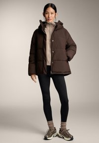Nainen, jolla on päällään ruskea untuvaparka, beige vetoketjullinen neule, mustat leggingsit, beiget sukat ja ruskeat lenkkarit, seisoo neutraalia taustaa vasten.