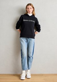 Schwarzer Kapuzenpulli mit weißem "Marc O'Polo"-Logo, kombiniert mit hellblauen Jeans, die dunklere blaue Seitenpaneele haben, und weißen Sneakers.