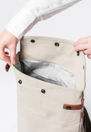 Hände, die einen geöffneten beigen Rucksack mit einem innen schwarz-weiß karierten Futter und schwarzen Druckknöpfen an der Klappe vor weißem Hintergrund halten.