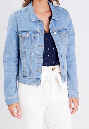 Femme portant une veste en denim bleu clair par-dessus un chemisier bleu marine avec de petits motifs, associée à un pantalon blanc taille haute noué avec une ceinture en tissu.
