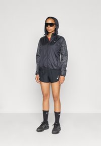 Zwarte windbreakerjack met mesh-mouwen, gecombineerd met zwarte shorts. Zwarte sportschoenen en crewlengte sokken. Donkere zonnebril gedragen.