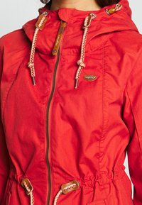 Veste d'extérieur rouge avec fermeture éclair, capuche à cordon de serrage et taille élastique. Comprend des accents en cuir et des cordons beiges tressés.