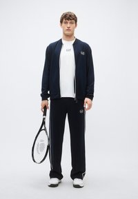 Tmavomodrá tepláková súprava s bundou na zips a zodpovedajúcimi nohavicami, na hrudi a bokoch s logom. Pod tým biele tričko, drží tenisovú raketu.