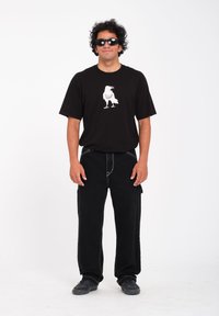 Zwarte t-shirt met een witte afbeelding van een raaf, gecombineerd met zwarte cargobroeken en zwarte sneakers. De outfit heeft een casual pasvorm.