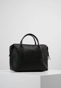 Svart texturerad läderhandväska med dubbla handtag och dragkedjestängning, placerad på en vit yta mot en enfärgad grå bakgrund.