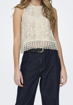 Jeune femme portant un haut en dentelle crème sans manches avec ourlet festonné et un jean bleu foncé taille haute avec une ceinture noire.