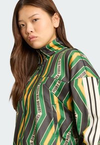 Veste zippée avec un motif géométrique vert, jaune et noir, col montant et bandes blanches contrastées sur les manches.