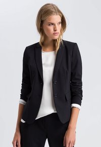Black blazer with a tailored fit, single-breasted design, two buttons, and two front pockets. White blouse underneath. Smooth fabric texture.