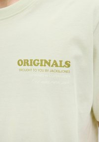 Lichtgroene katoenen t-shirt met vetgedrukte tekst "ORIGINALS" in olijfgroen, met daaronder extra tekst in een kleinere, lichtere lettertype. Korte mouwen.