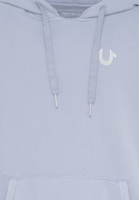 Sudadera azul claro con bolsillo canguro, cordones y un pequeño logo blanco en el pecho. La tela es suave y ligeramente texturizada.