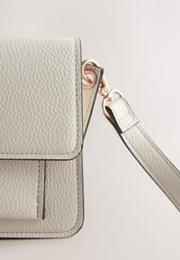 Sac à main en cuir texturé gris clair avec une bandoulière amovible attachée par des fermoirs en or rose sur un fond uni.