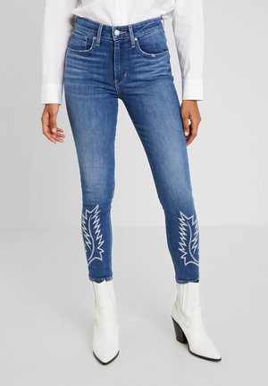 Femme portant un jean skinny bleu avec des motifs brodés blancs sur les mollets, une chemise blanche boutonnée et des bottines blanches à talons.