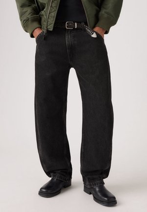 Personne portant un jean noir à jambes larges, des bottes en cuir noires, une ceinture tissée noire et une veste verte avec les mains dans les poches.