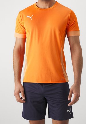 Mężczyzna w jaskrawo pomarańczowej sportowej koszulce z krótkim rękawem z białym logo Puma i granatowych szortach Puma na jednolitym jasnym tle.