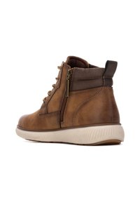 Botín de cuero marrón hasta el tobillo con una superficie texturizada, cuello acolchado, cremallera lateral y una suela de goma beige con surcos horizontales.