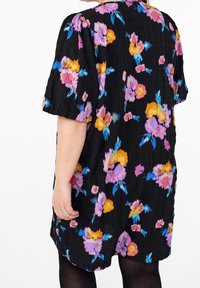 Femme portant une robe noire à motifs floraux violets, roses, jaunes et bleus, vue de dos, avec des manches courtes et un tissu texturé.
