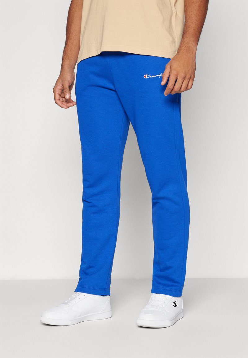 Blå joggebukser med en myk tekstur, elastisk midje, og en liten hvit logo. Kombinert med hvite sneakers og en beige skjorte.