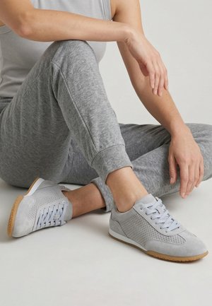 Persoon zit met gekruiste benen, draagt lichtgrijze sneakers, bijpassende grijze joggingbroek en een mouwloos topje tegen een effen achtergrond.