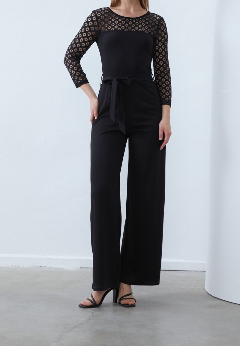 Femme portant une combinaison noire avec manches en dentelle, une taille ceinturée et un pantalon à jambes larges, debout sur un sol en béton en talons.