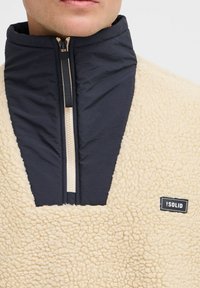 Fleece pullover with a high collar, cream textured fabric and dark blue nylon panel, features a central zipper and a black brand label.