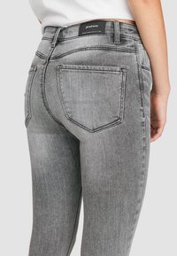 Jeans de denim gris con un diseño ajustado, que cuentan con bolsillos traseros y una etiqueta en la cinturilla. Costura distintiva a lo largo de las costuras.