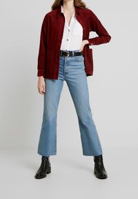 Rode corduroyen overhemdjas, witte blouse, lichtblauwe flare jeans, zwarte leren enkellaarzen, met een zwarte riemdetails.