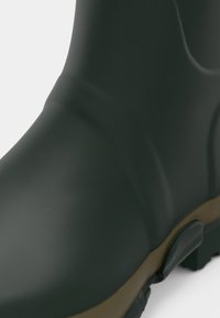 Πράσινη αδιάβροχη μπότες με ματ φινίρισμα, υφή στην άνω επιφάνεια και καφέ καουτσούκ σόλα που διαθέτει σχέδιο ανάγλυφης πρόσφυσης.