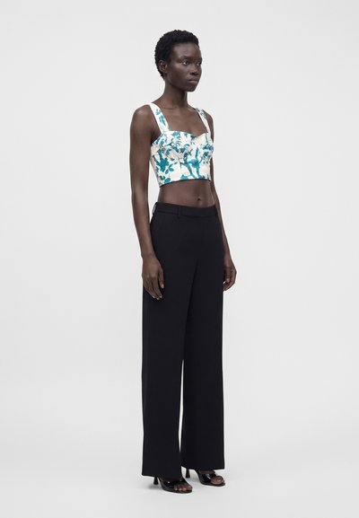 Femme portant un crop-top fleuri bleu et blanc avec de larges bretelles, un pantalon noir taille haute et jambes larges, ainsi que des talons noirs ouverts, debout de profil.
