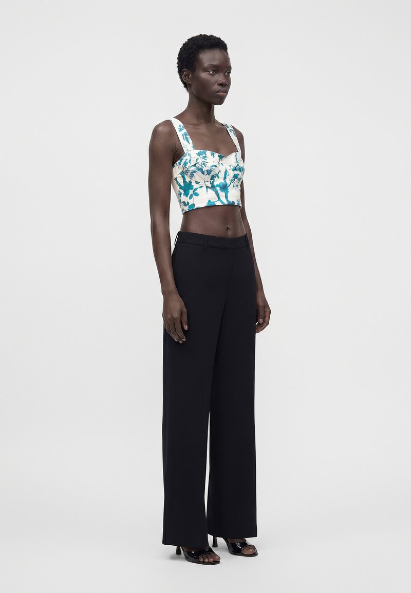 Woman wearing a blue and white floral crop top with wide straps, black high-waist wide-leg trousers, and black open-toe heels standing sideways.